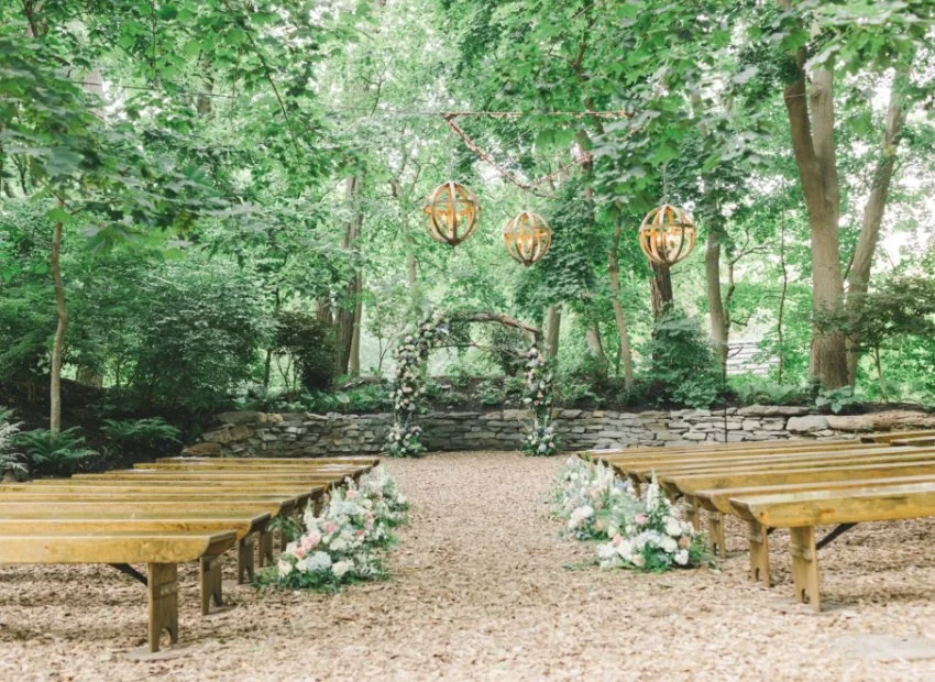 Outdoor wedding ceremony setup in a wooded area with wooden benches, floral decorations, and hanging lanterns.
