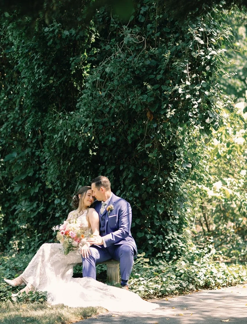 A bride and groom sitting close together outdoors, surrounded by lush greenery. The bride holds a bouquet of flowers, and both are dressed in wedding attire, sharing an intimate moment.
