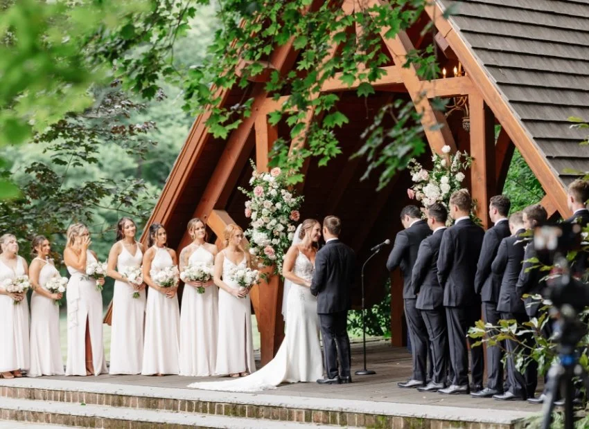 Wedding ceremony with bride and groom standing under a wooden arch decorated with flowers, surrounded by bridesmaids and groomsmen outdoors.