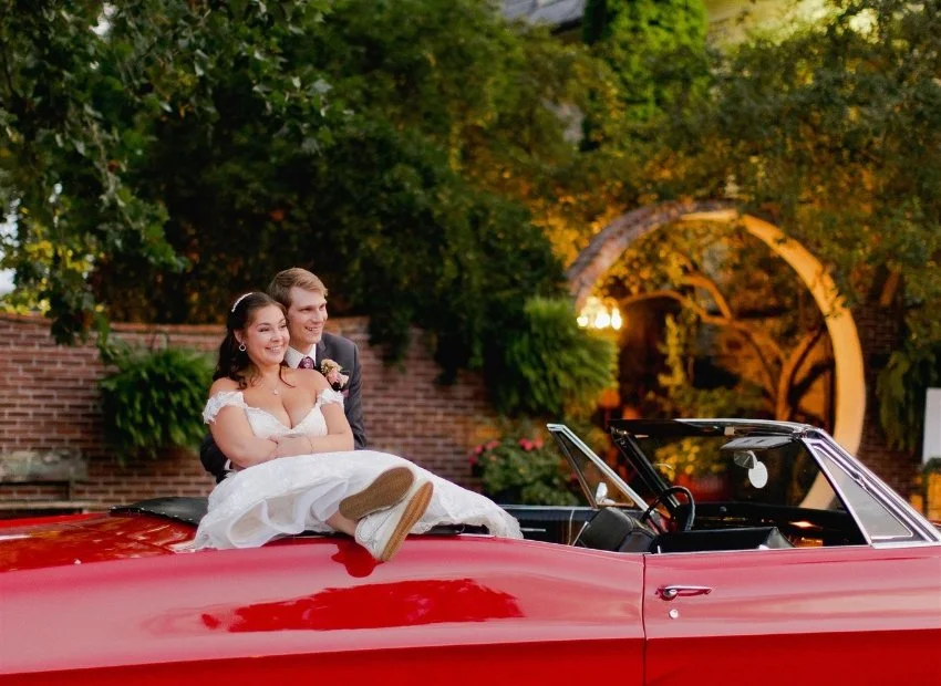 Happy bride and groom sitting together on the back of a red convertible car in an outdoor setting with green trees, brick wall, and colorful lighting in the background.