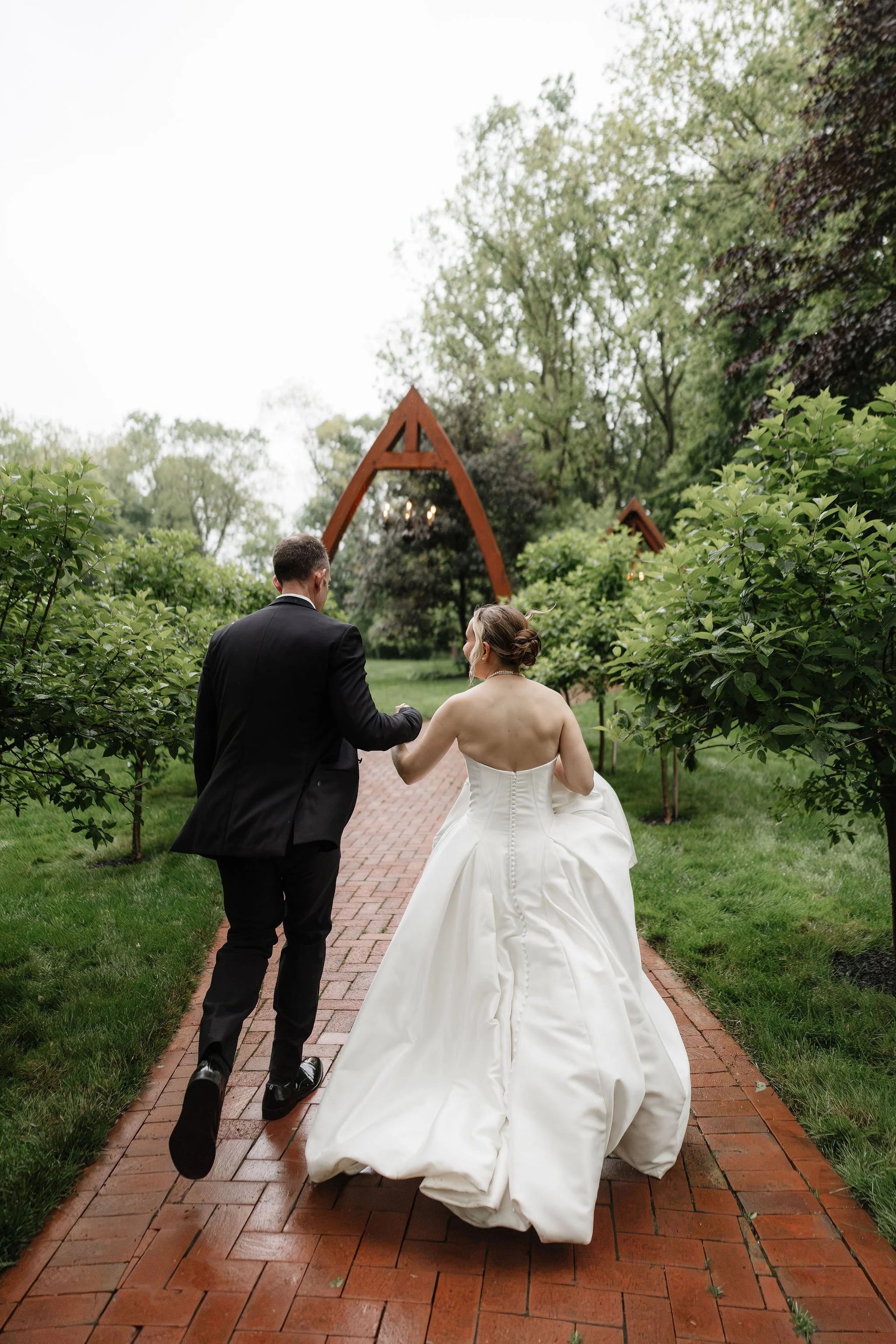 A bride and groom walking hand in hand on a brick path surrounded by lush green trees and bushes, with a wooden arch structure in the background during daytime.