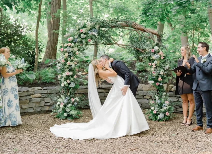 A bride and groom sharing a kiss during their outdoor wedding ceremony, with floral arch and guests clapping.