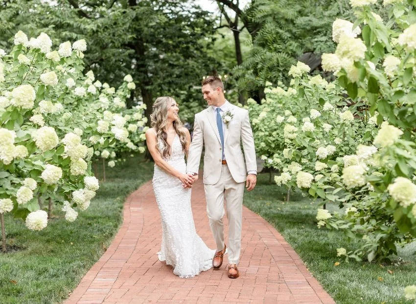 A bride and groom walking hand in hand on a brick path surrounded by white hydrangea bushes.