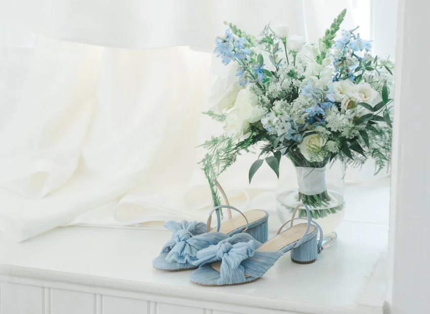 A bouquet of white and light blue flowers in a glass vase next to a pair of light blue high-heeled shoes decorated with bows.