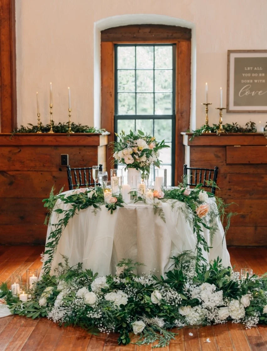 A decorated table with a floral centerpiece, surrounded by candles and greenery, in front of a window with wooden trim and a framed wall art piece on the right, in a rustic indoor setting.