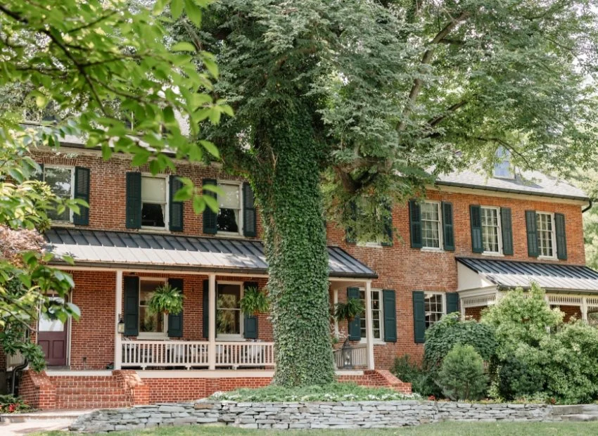 A large brick house with black shutters and multiple windows, surrounded by lush green trees and bushes, with a stone wall in the front yard.