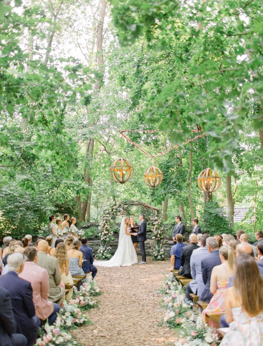 A wedding ceremony taking place outdoors in a lush, green forest, with a bride and groom exchanging vows under decorative floral arch, surrounded by seated guests, with hanging lanterns above.