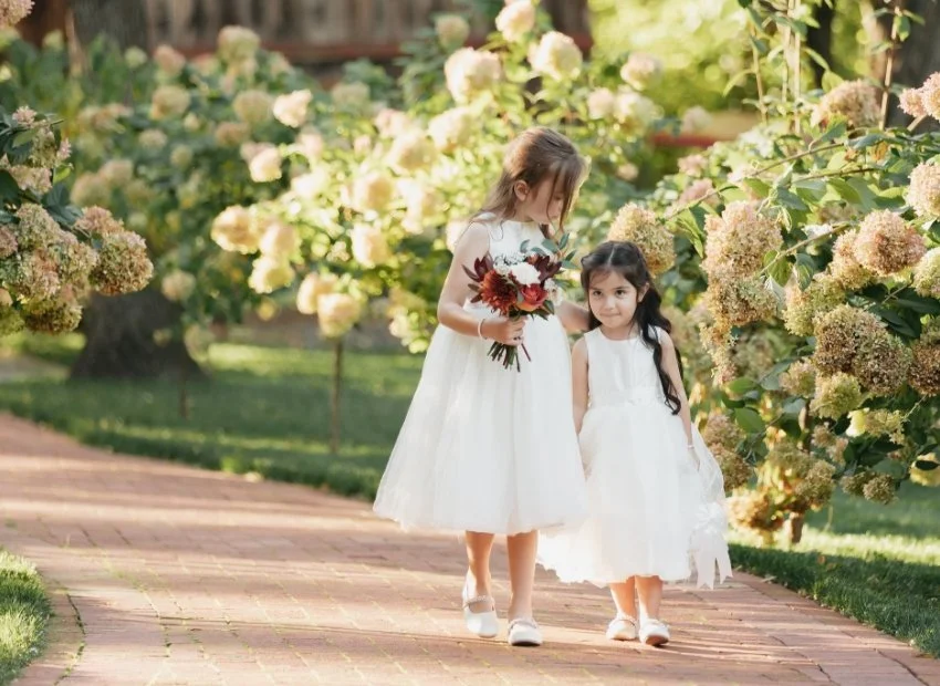 Two young girls in white dresses walking on a brick path, surrounded by blooming hydrangea bushes, with one girl holding a bouquet of flowers.
