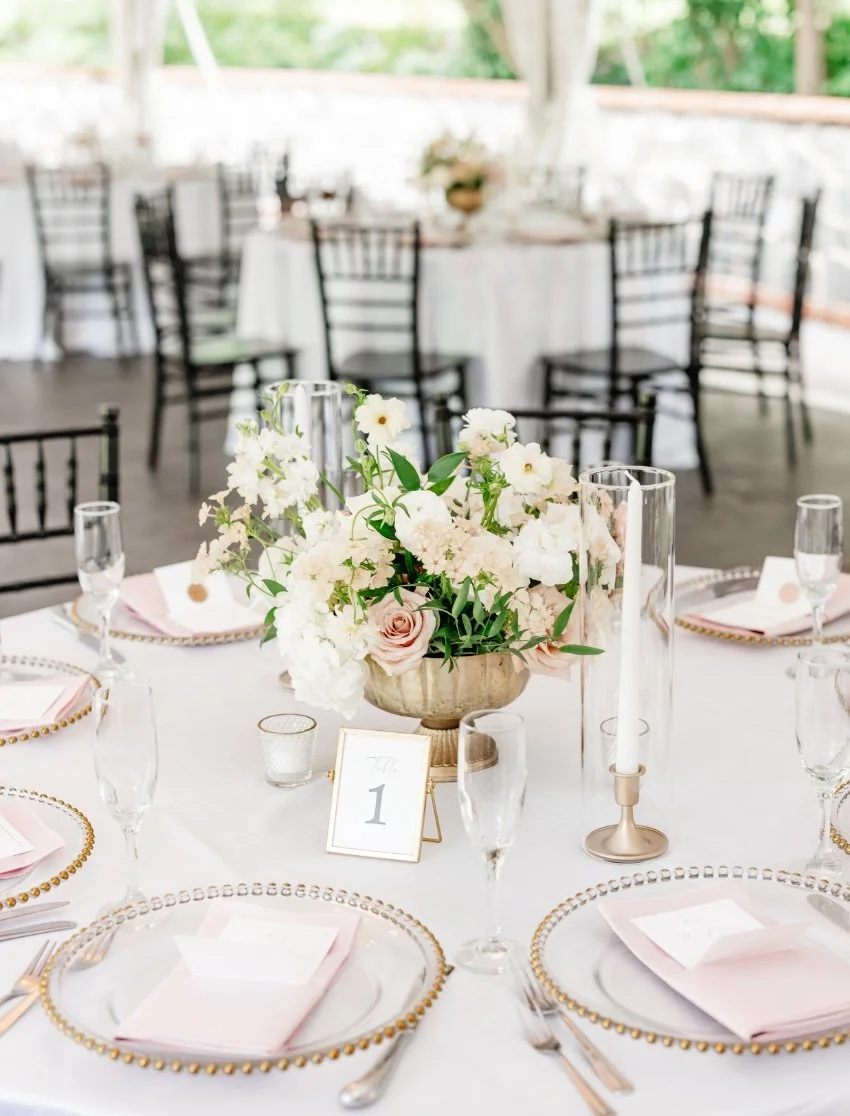 Elegant wedding reception table decorated with a floral centerpiece, pink napkins, and gold-rimmed glass chargers.
