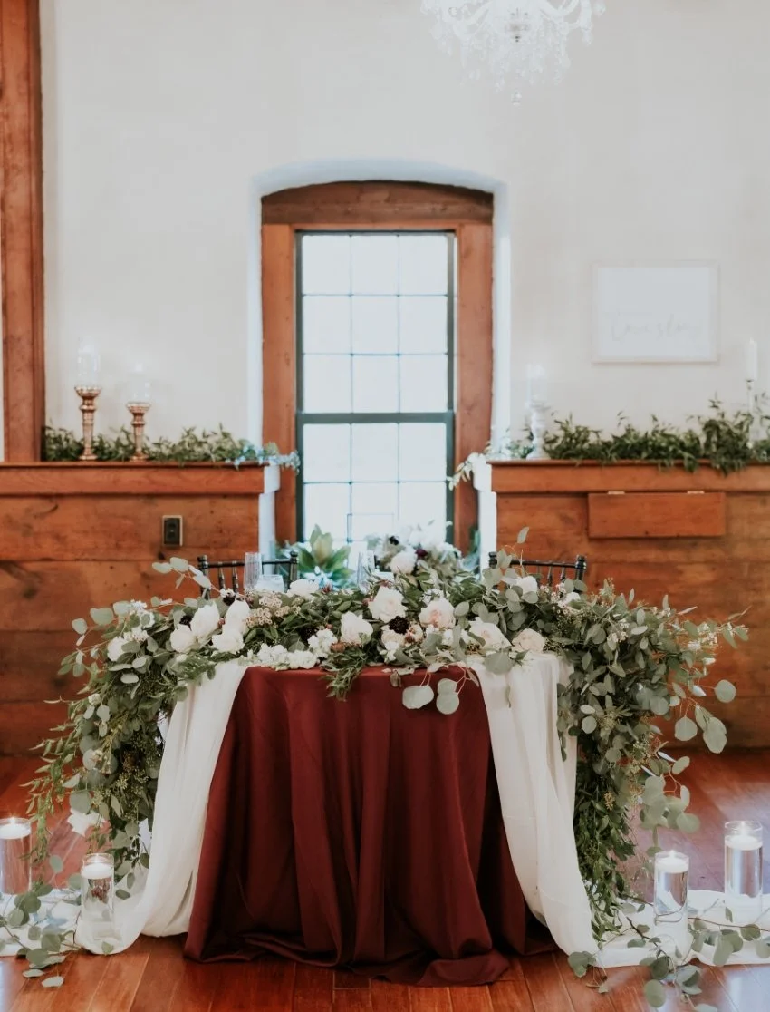 A wedding or event table decorated with a floral centerpiece and draped fabric, set against a wood-paneled wall with candles and greenery, in a rustic-style room with a large window.