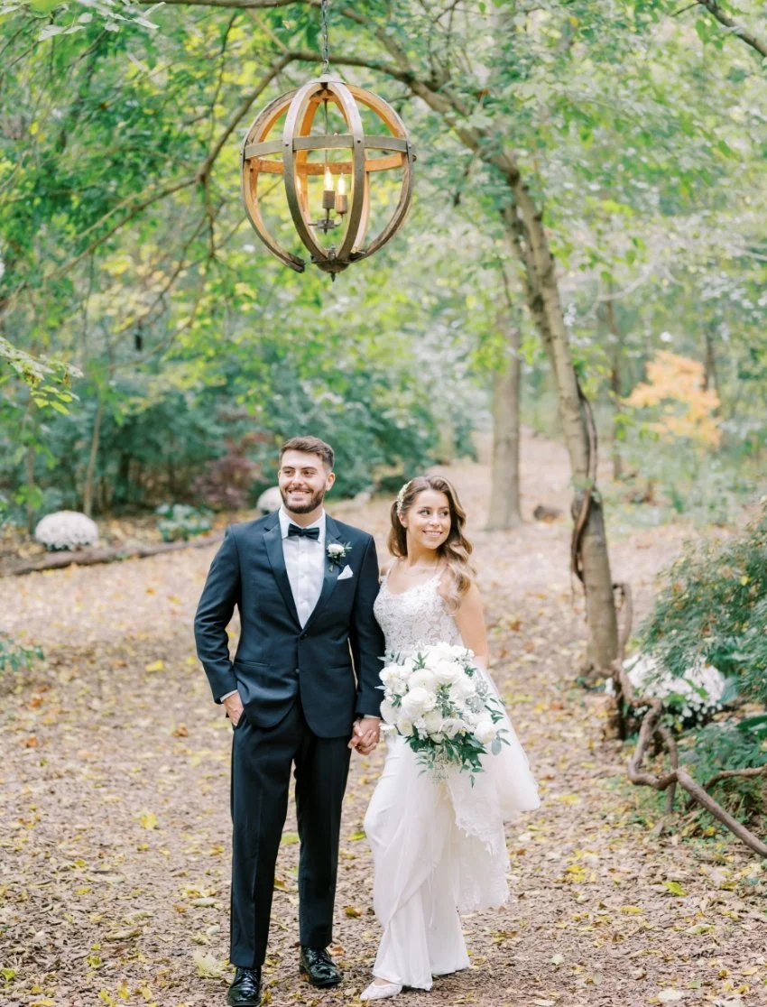 Bride and groom holding hands and walking outdoors in a wooded area, dressed in wedding attire, with a wooden chandelier hanging overhead.