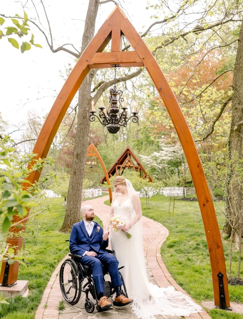 A bride and groom, with the groom in a wheelchair, stand together outdoors on a brick pathway surrounded by green grass and trees. The bride holds a bouquet of flowers and wears a white wedding dress with a veil. A decorative black chandelier hangs f