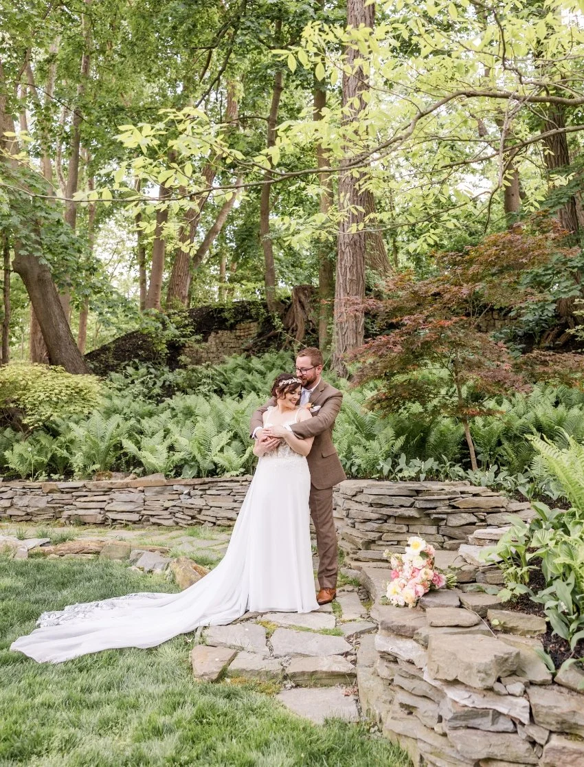 A bride and groom standing close together on a stone pathway amidst lush green foliage and trees in a garden setting, with a bouquet of flowers placed on the rocks nearby.