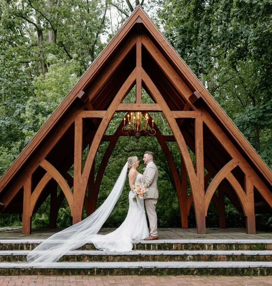 A bride and groom standing under a wooden pavilion with a triangular roof, holding hands and facing each other, with a hanging chandelier above and trees in the background.