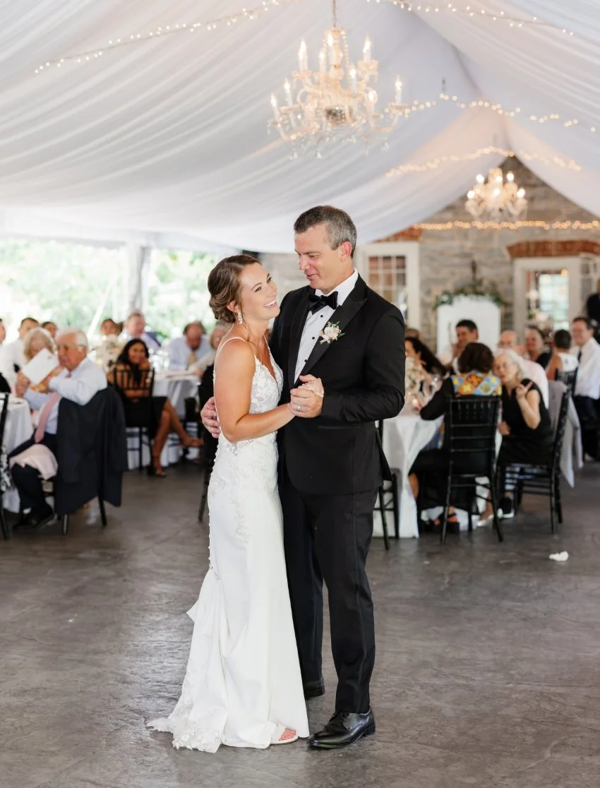 A bride and groom dancing at their wedding reception, surrounded by seated guests under a decorated tent with chandeliers and string lights.