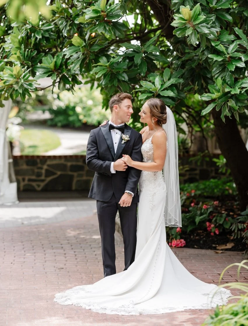 A bride and groom in wedding attire standing outdoors under a tree, holding hands and looking at each other with affectionate expressions.