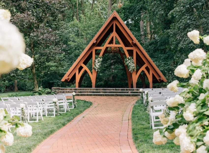 A wedding ceremony setup outdoors featuring a brick aisle leading to a wooden A-frame structure with floral decorations, surrounded by green trees and white chairs with floral accents.