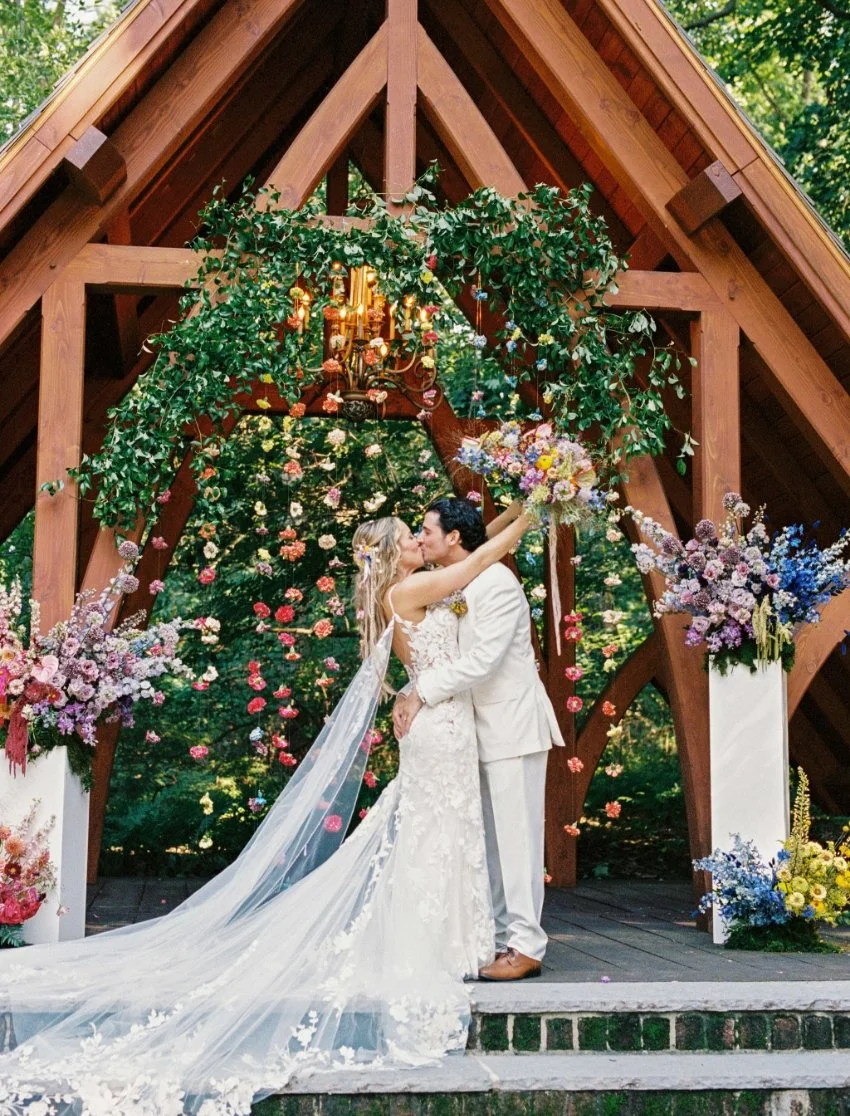 A bride and groom kiss on a wedding altar decorated with flowers and greenery, surrounded by a wooden structure and a large outdoor floral display.
