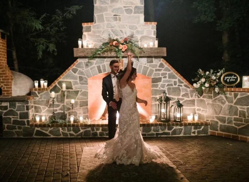 A bride and groom are dancing in front of a decorated firepit at night, surrounded by candles and floral arrangements.