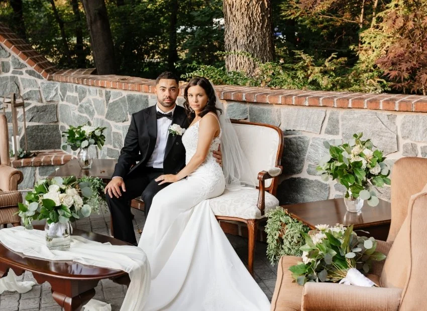 A bride and groom sitting on a vintage sofa outdoors, with greenery and trees in the background. The bride wears a white wedding dress with lace details and a veil, while the groom is in a black tuxedo with a bow tie. They are surrounded by floral ar
