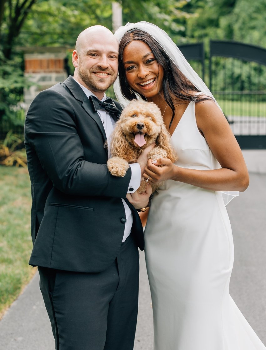 A happily married couple in wedding attire posing outdoors with a small, fluffy dog. The groom is in a black tuxedo, and the bride is in a white wedding dress and veil. They are holding the dog, who has its tongue out, and are smiling at the camera.