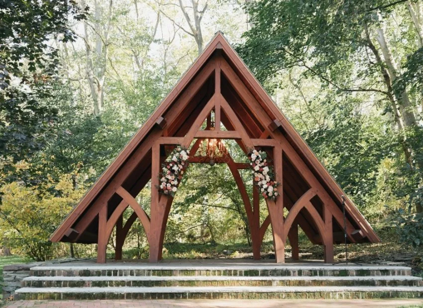 A wooden wedding arch decorated with pink and white flowers in an outdoor forest setting with trees.