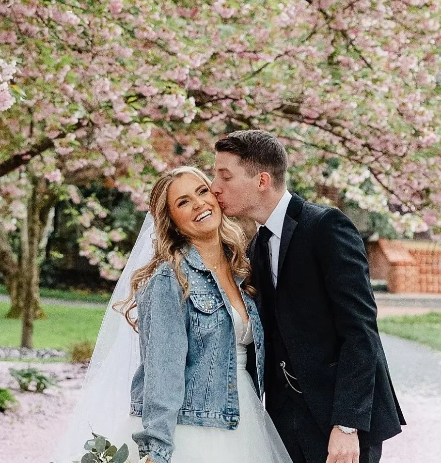 A couple dressed for a wedding, with the man kissing the woman's cheek, standing under pink blossoming trees in a park or garden setting.