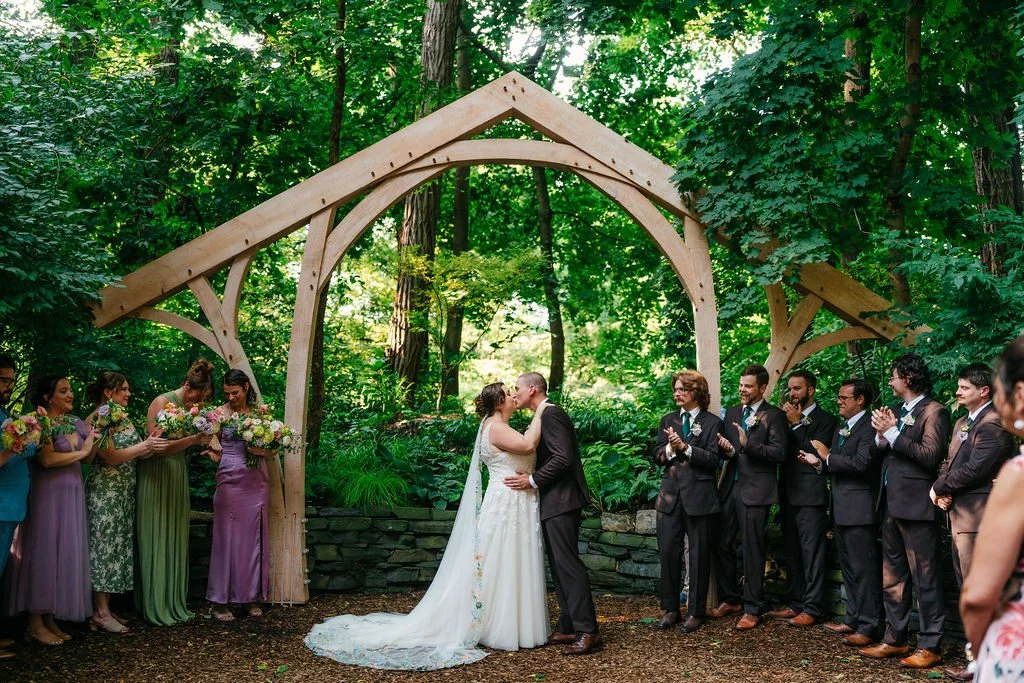 Couple kissing at their outdoor wedding ceremony under a wooden arch, surrounded by wedding party and guests in a lush green forest setting.