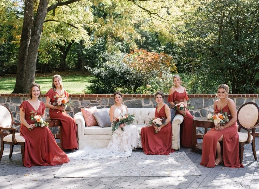 Bridal party poses outdoors on a shaded patio with a brick wall and trees in the background. The bride, in a white gown, sits on a white sofa, surrounded by five bridesmaids in matching rust-colored dresses, each holding bouquets. The scene is bright