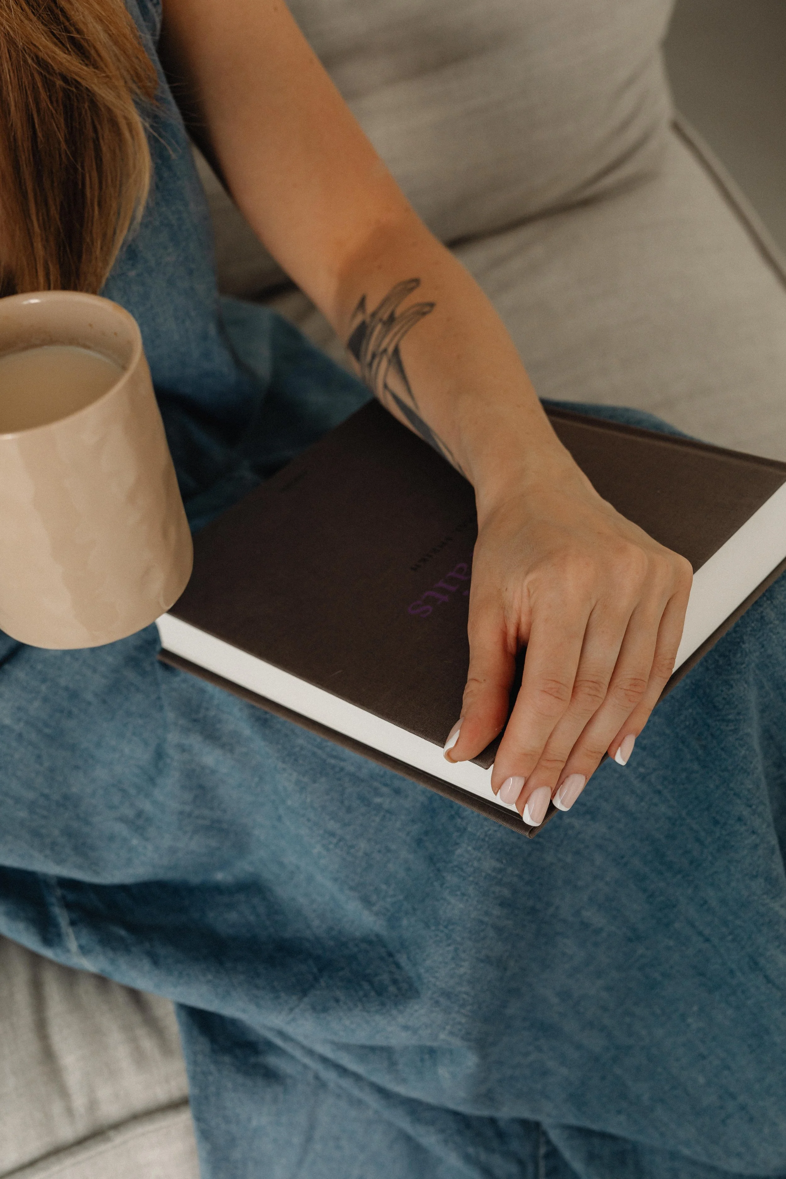 Person sitting on a sofa, holding a coffee mug in one hand and a book in the other, with a visible tattoo on their forearm.