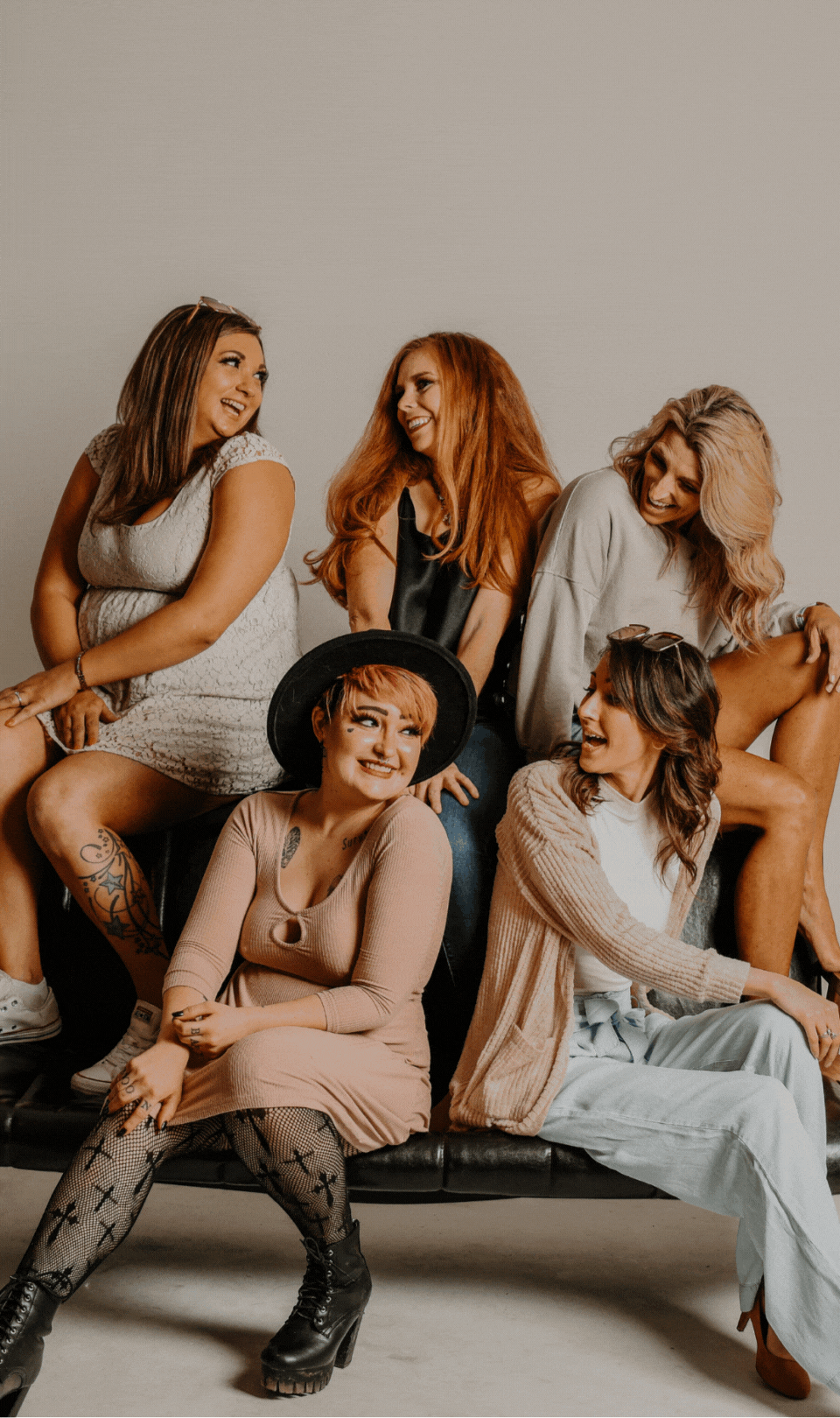 Six women sitting and smiling together, dressed in casual and trendy clothing, against a plain background.