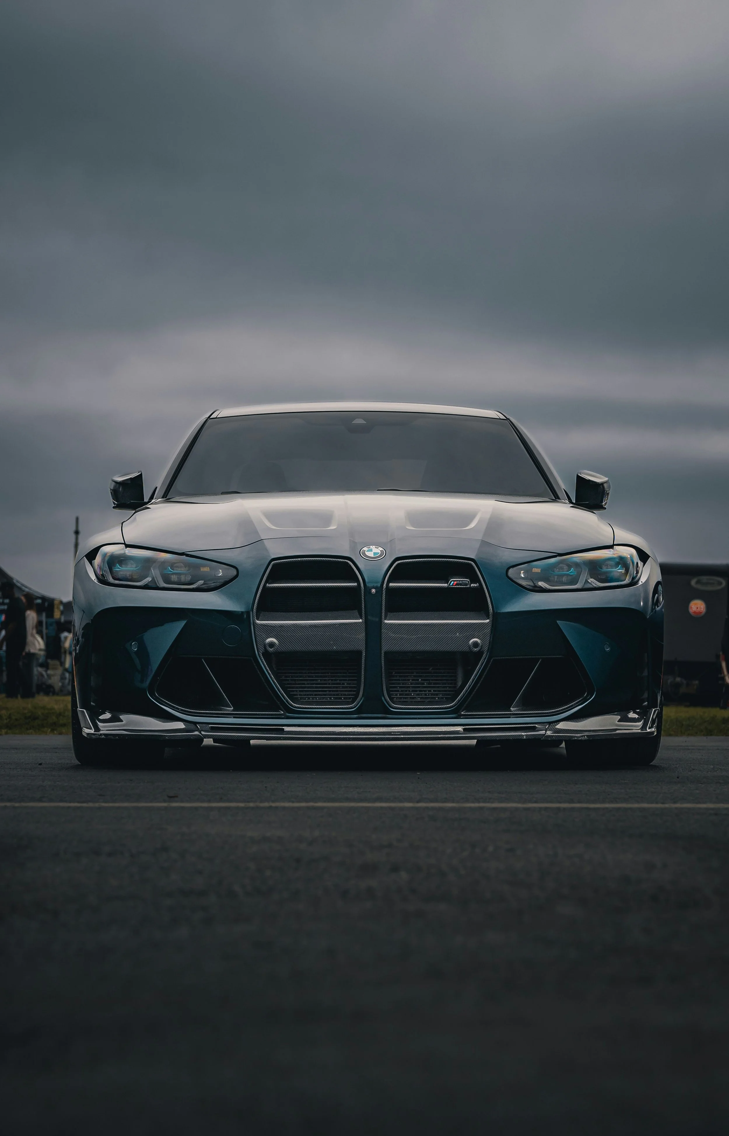 A black BMW car with a large front grille on a cloudy day, parked on a street or track.