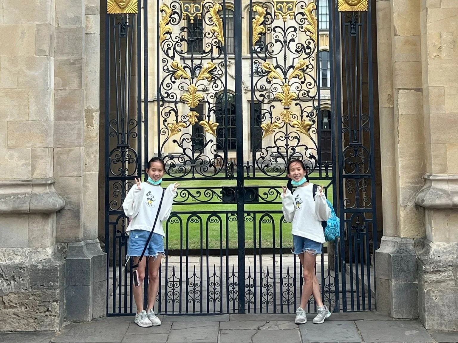Two young women standing in front of an ornate black and gold iron gate, smiling and making peace signs. Behind the gate is a historic building with large windows and a green lawn.