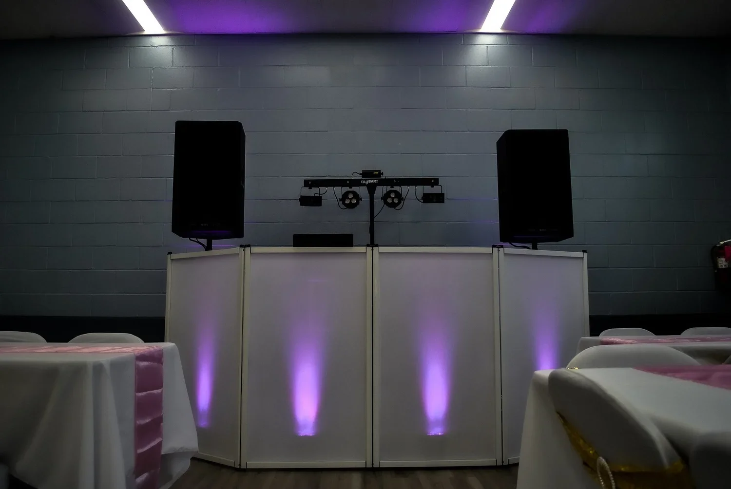 Empty event space with a DJ booth, large black speakers, lighting equipment, and tables with white tablecloths and pink accents, illuminated by purple lighting.