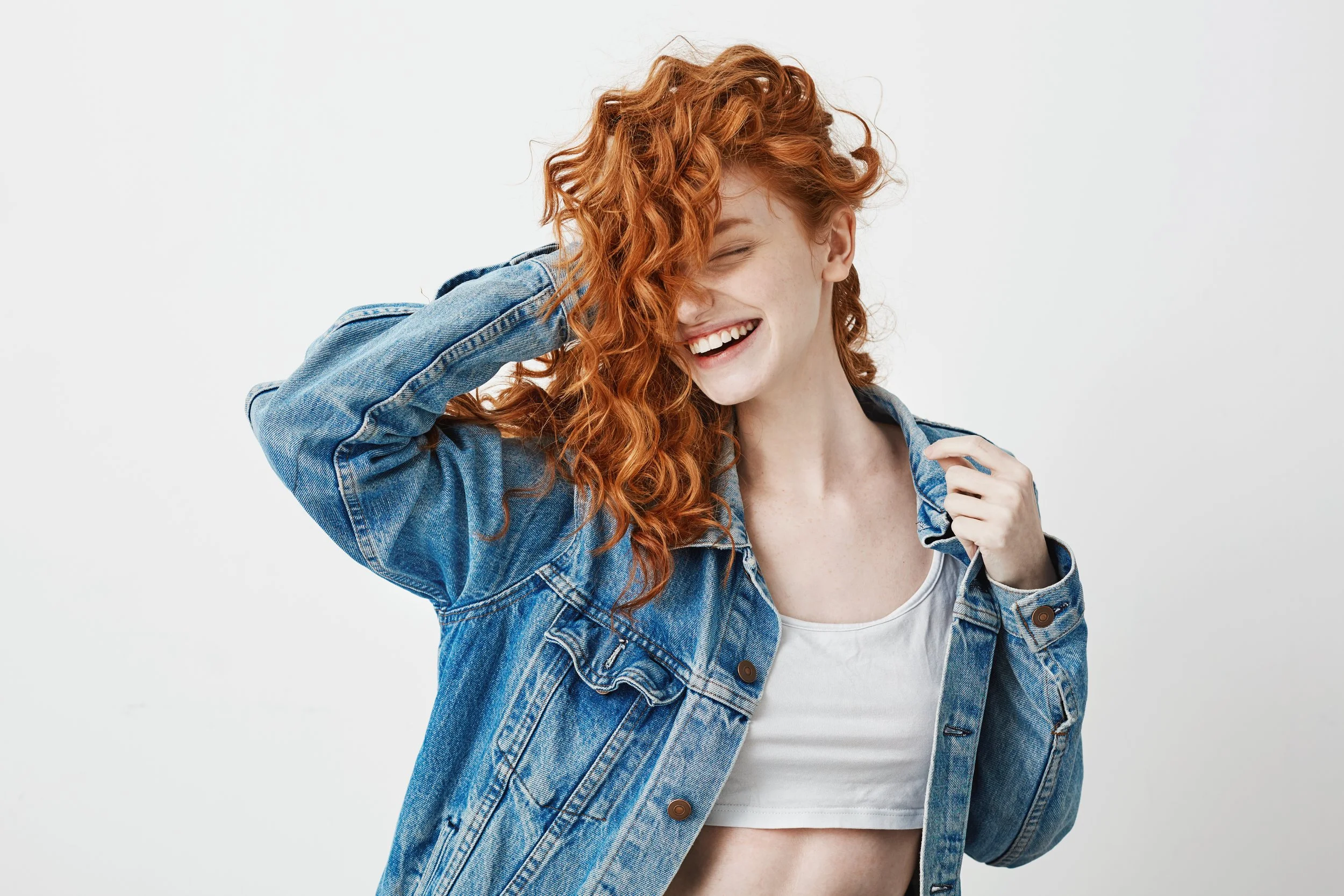 A young woman with curly red hair, smiling and laughing, wearing a denim jacket over a white crop top, standing against a plain white background.