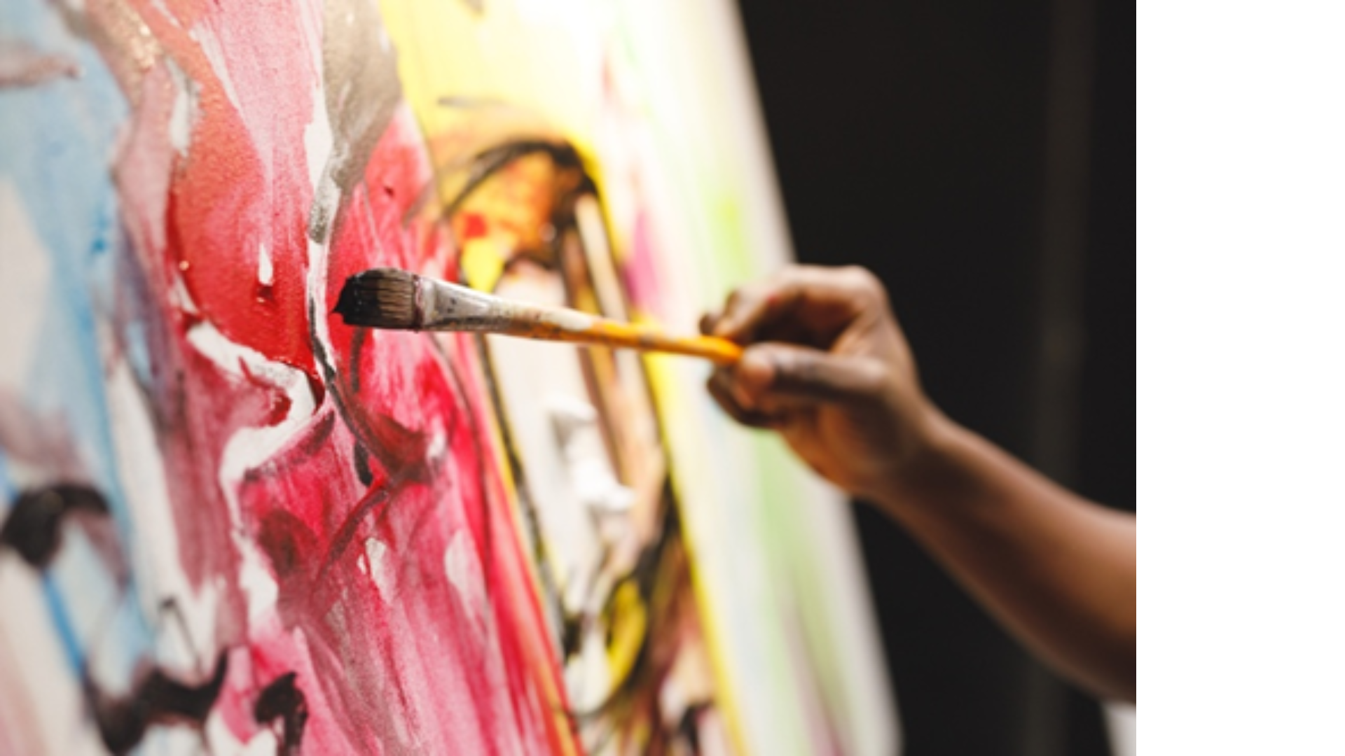 Close-up of an artist's hand holding a paintbrush, painting an abstract artwork with red, pink, white, yellow, and black colors on a canvas.