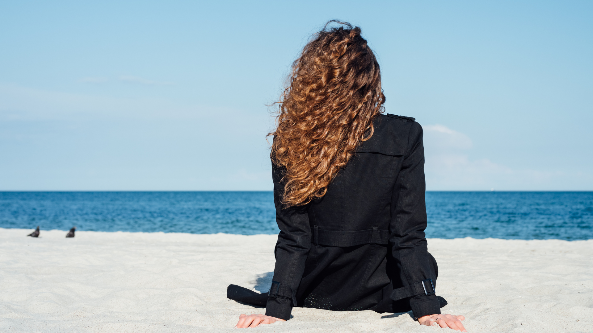 A woman with long curly hair wearing a black jacket sitting on a sandy beach, facing the ocean.
