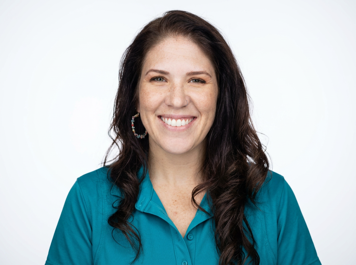 A woman with long dark wavy hair, light skin with freckles, smiling, wearing a teal collared shirt and colorful hoop earrings, against a plain white background.