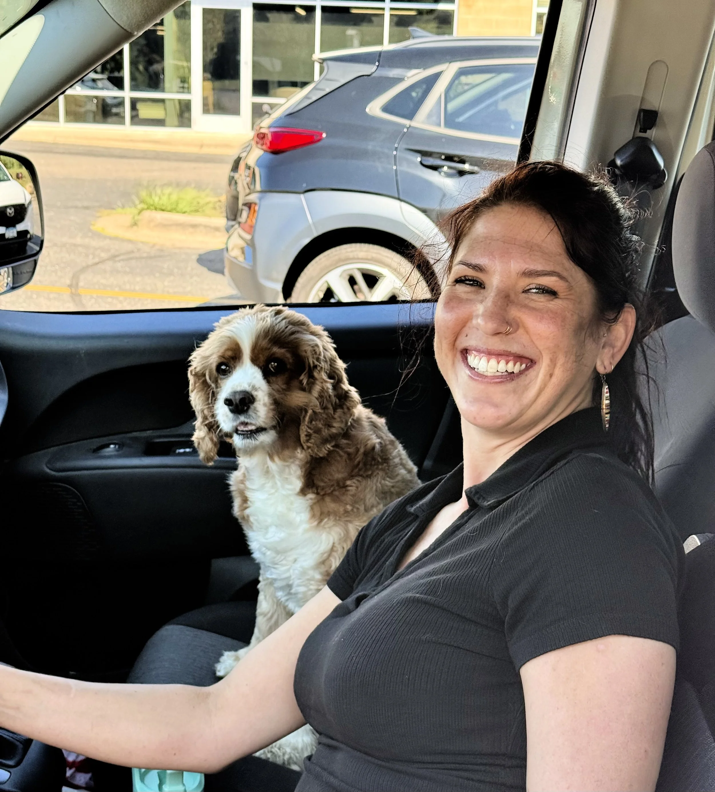 A woman smiling with a puppy on her lap inside a vehicle, with parked cars and a building with glass doors in the background.
