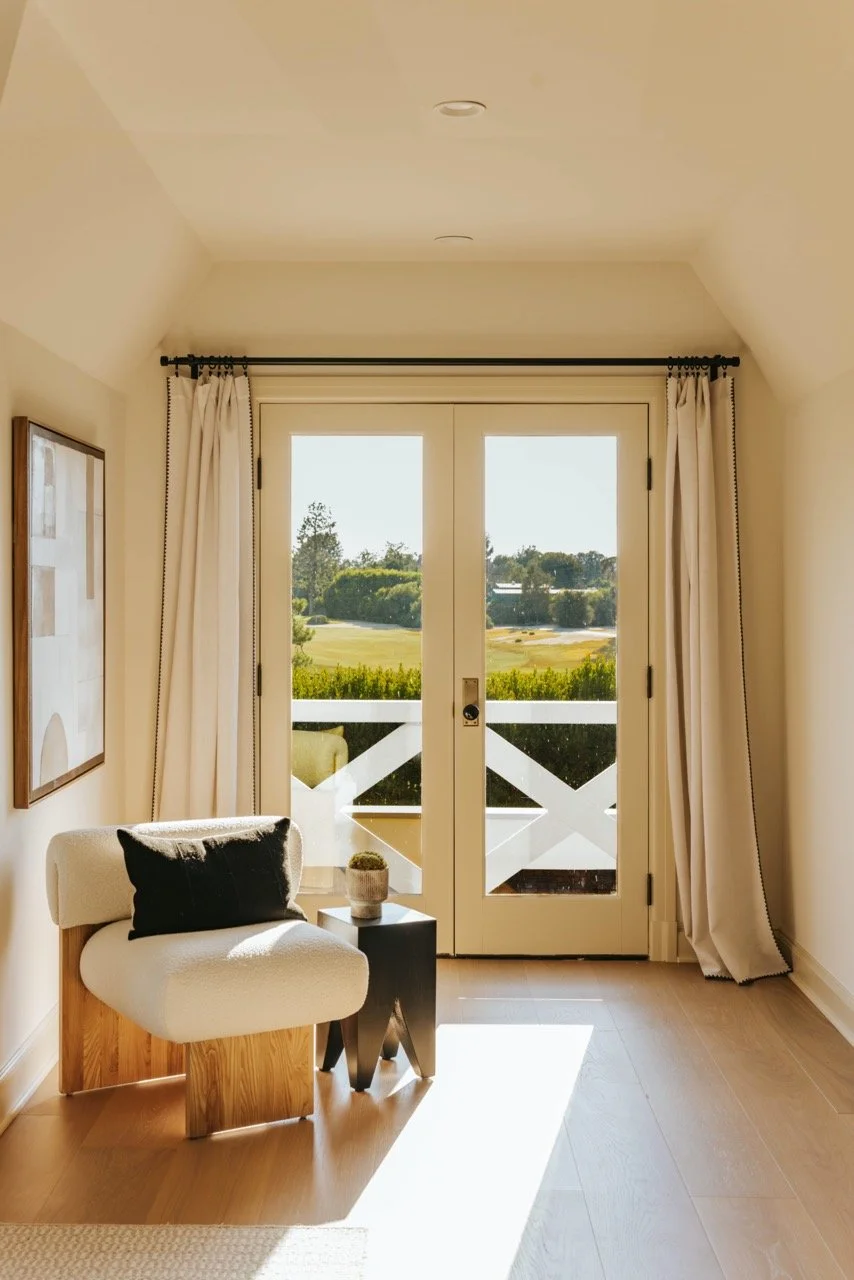Interior with modern chair, small side table, French doors with curtains, and outdoor garden view during daytime.