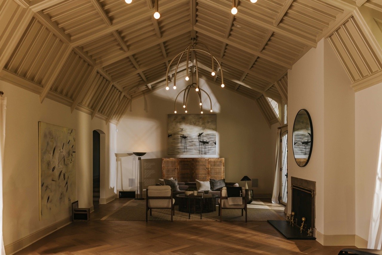 Luxurious living room with high vaulted ceiling, geometric chandelier, modern art on walls, hardwood flooring, fireplace, and stylish furniture arrangement.