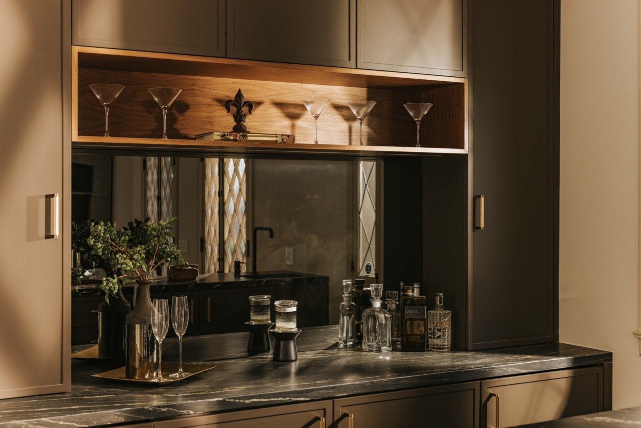 Modern kitchen with dark cabinetry, marble countertop, glassware on counter and shelves, decorative shelf with martini glasses, and greenery.