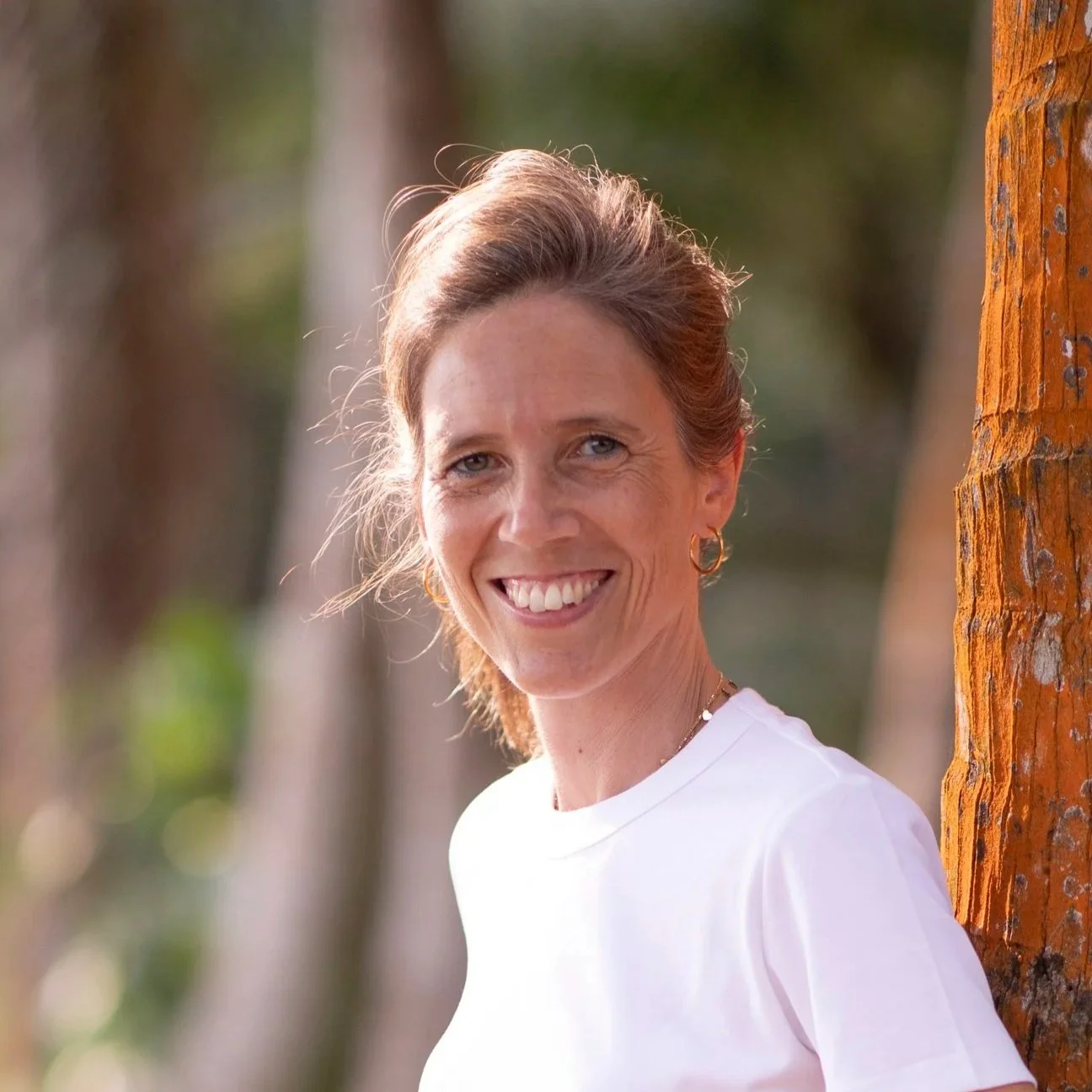 Une femme souriante avec des cheveux bruns attachés, portant des boucles d'oreilles en or, debout près d'un arbre dans un environnement naturel forestier.