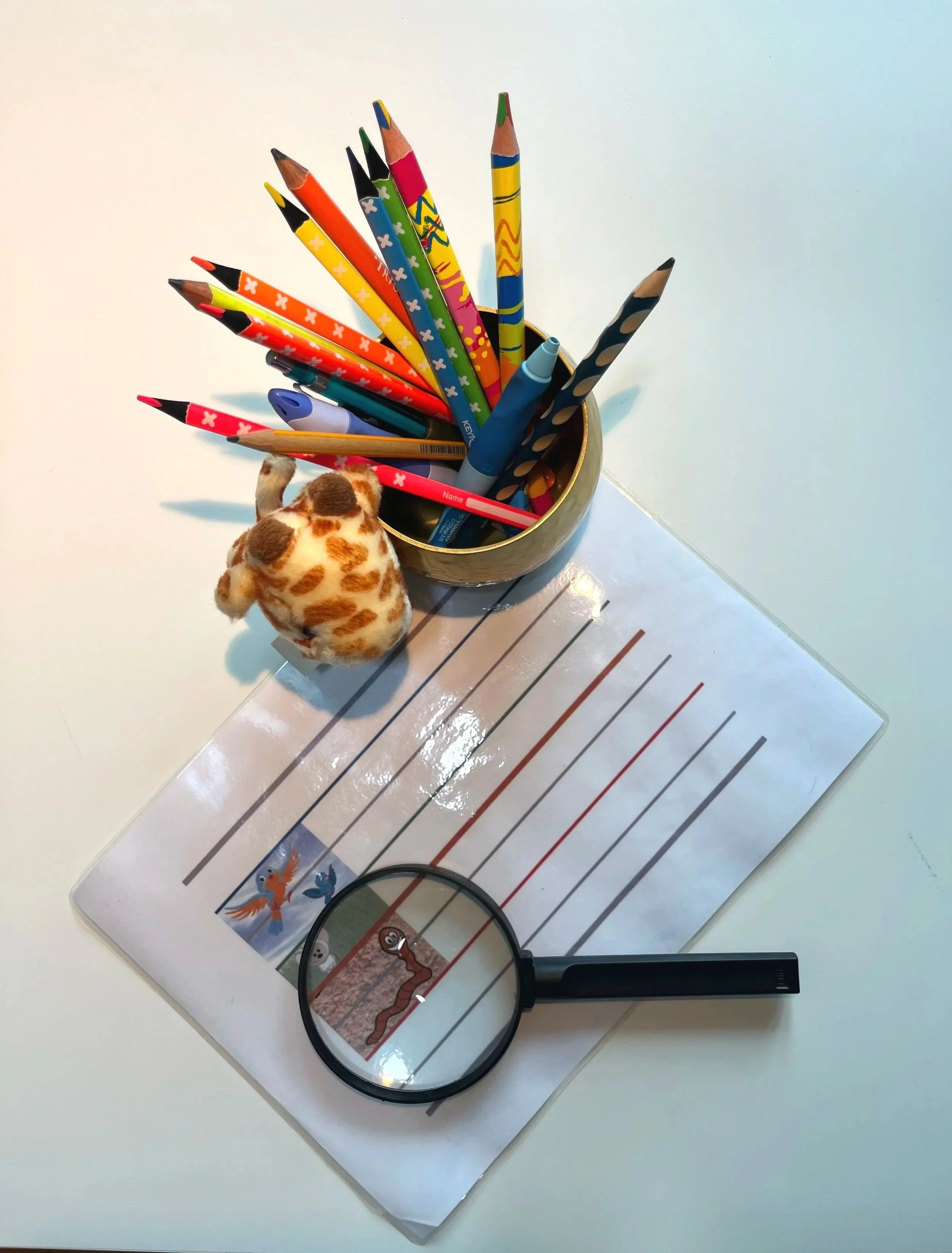 Un tableau avec des lignes colorées, une loupe, un pinceau de coloriage, une tasse remplie de crayons de couleur, et une petite peluche de girafe en peluche.