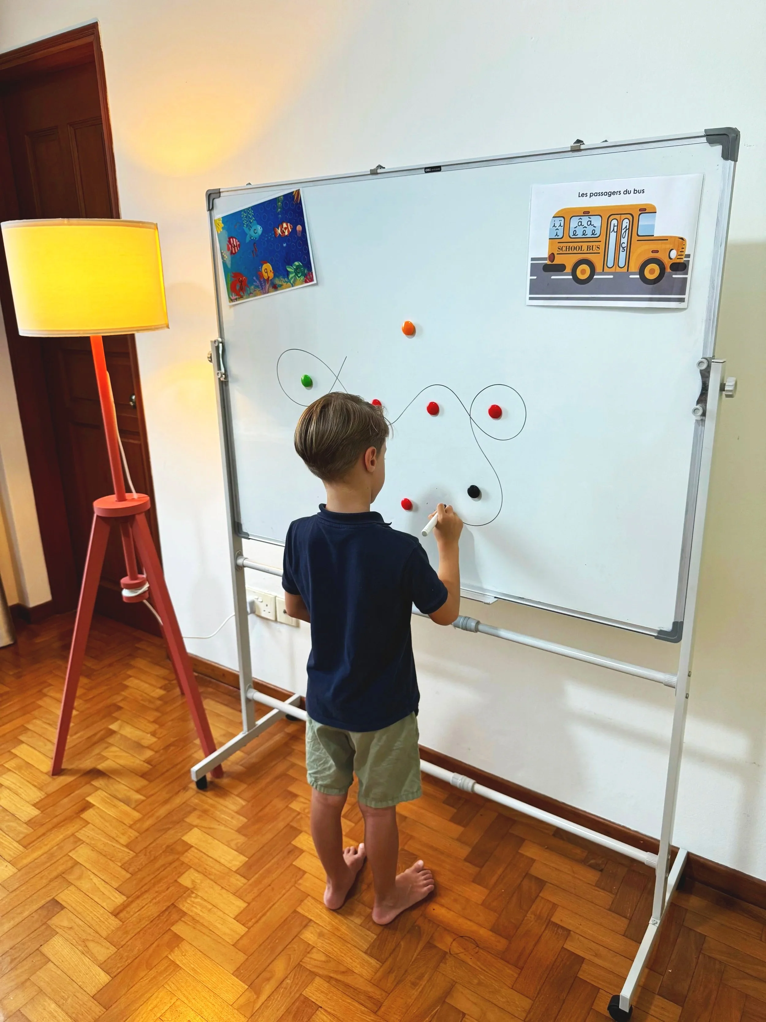 Un garçon dessine sur un tableau blanc avec un marqueur, avec deux images accrochées, une d'un bus scolaire et une d'un spectacle sous la mer, dans une pièce avec un sol en parquet et une lampe à pied. Il est pieds nus.