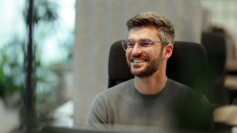 En mand med brunt hår, skæg og briller smiler og sidder i en kontorstol i et kontormiljø.