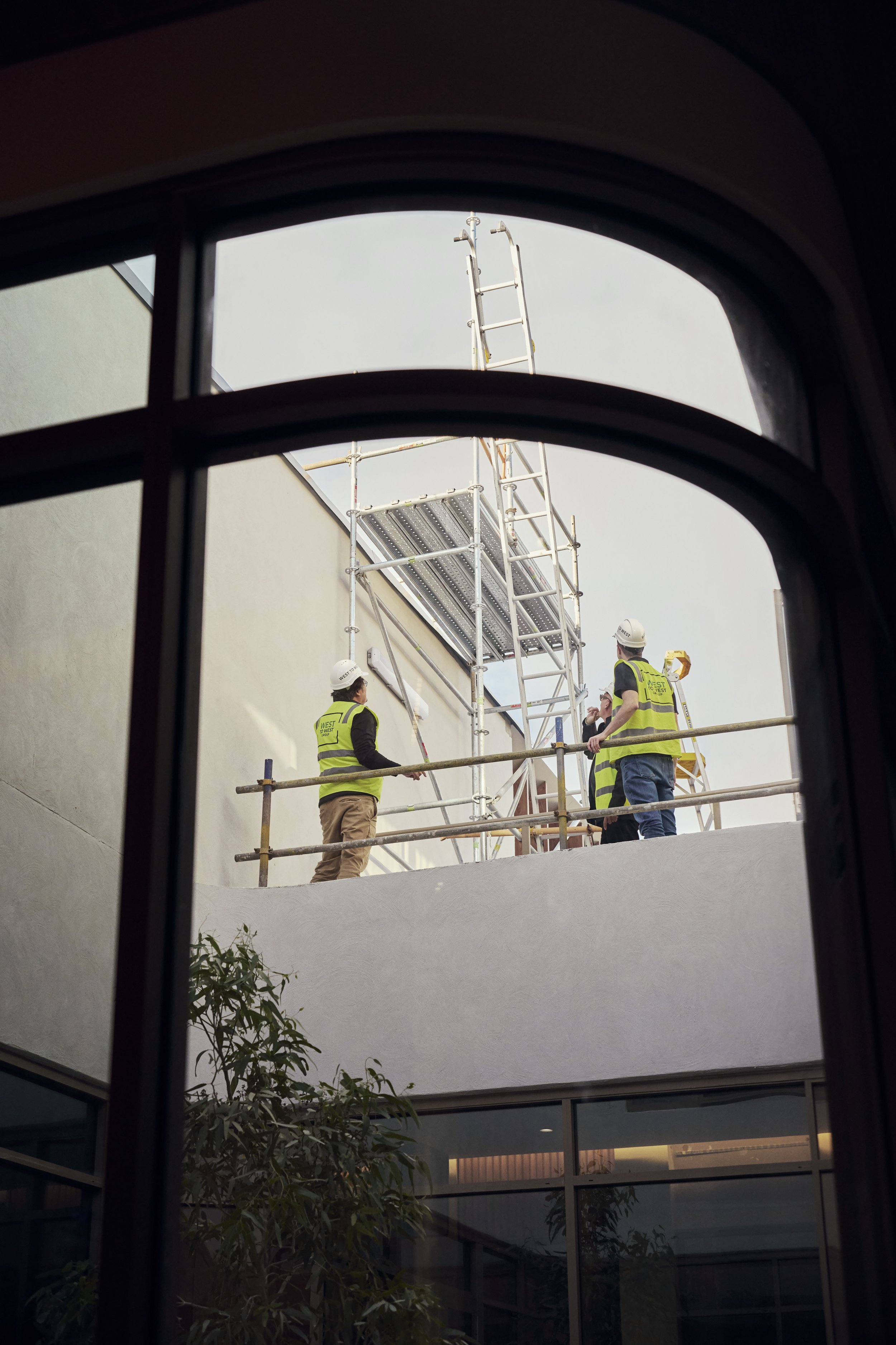 Construction workers in safety vests and helmets working on scaffolding outside a building, seen through a window.