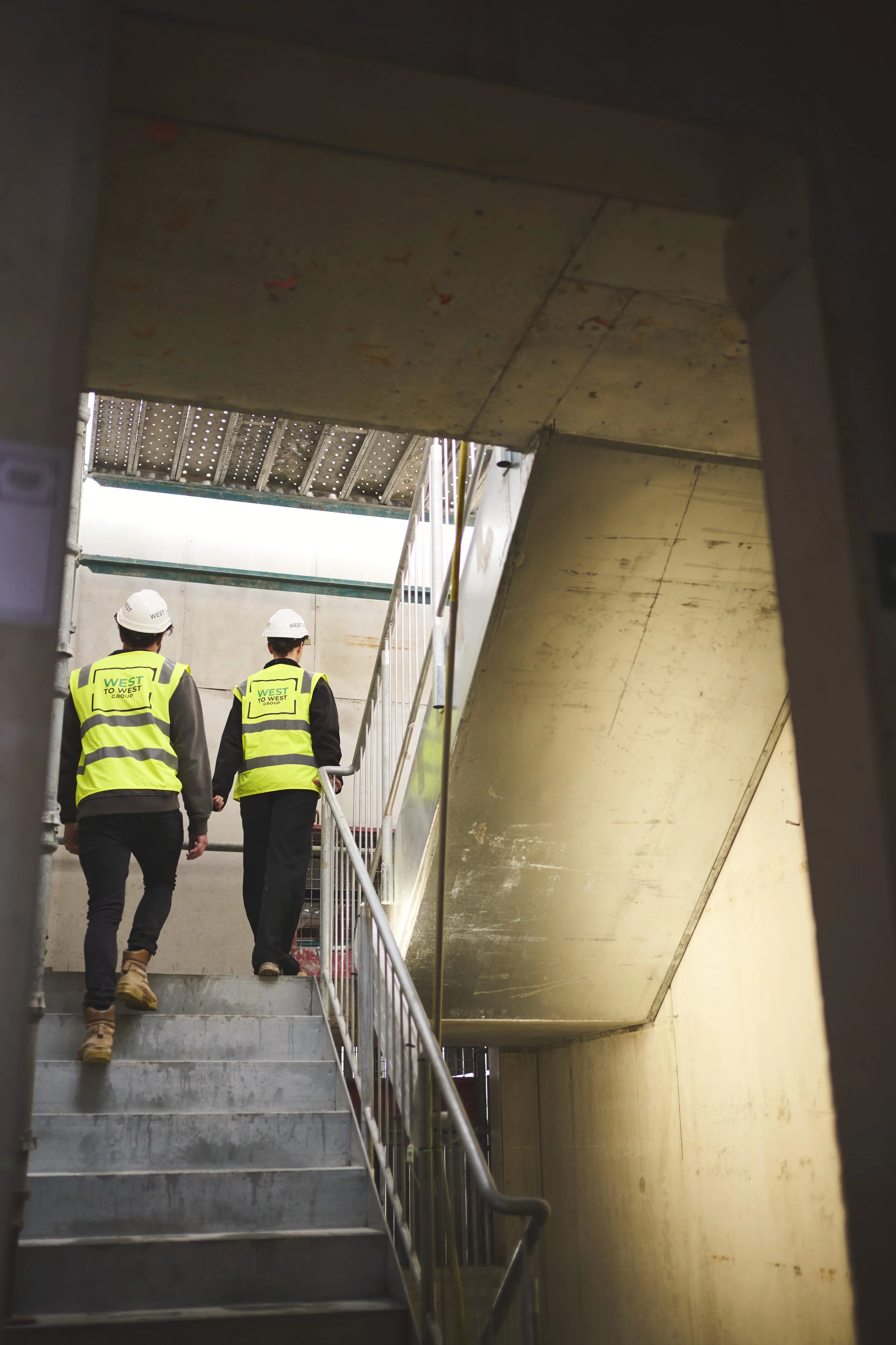 Two construction workers wearing yellow safety vests and white hard hats walking up stairs in an underpass or underground area with concrete walls and ceiling.