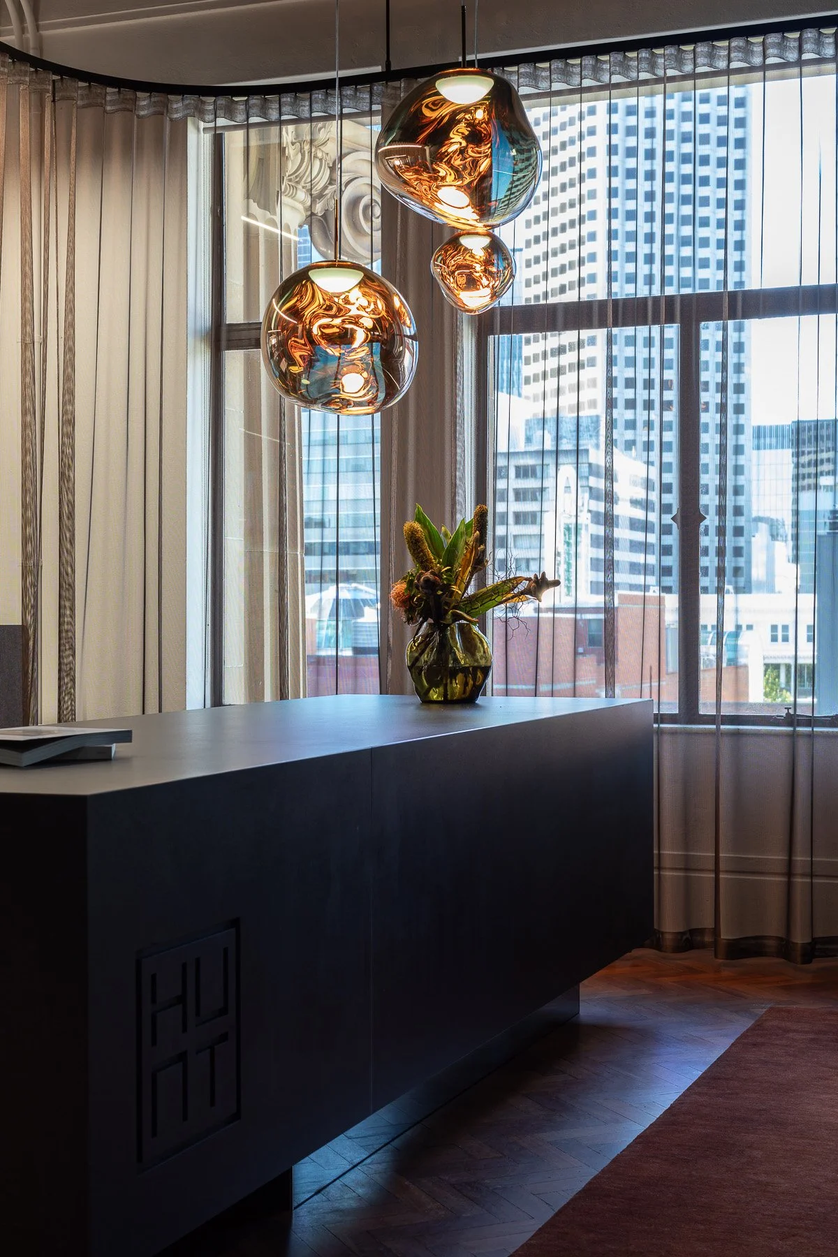Interior view of a modern office or lobby with black countertop, decorative vase with flowers, large window with cityscape, and three hanging metallic pendant lights.