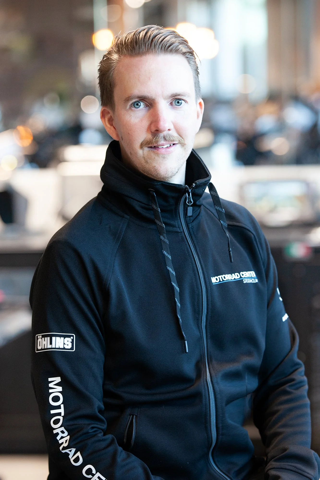 Man i svart jacka med "Motorrad Center"-logotypen, sitter i en lokal med motorcyklar i bakgrunden.